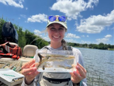 Emily Eberly holds a clear container containing several juvenile fishes to the camera.